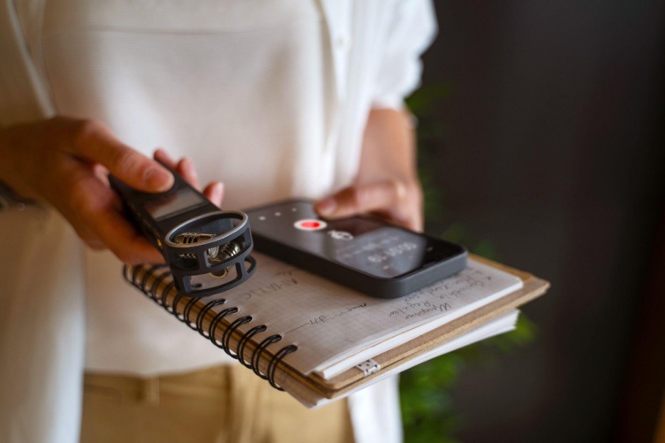 A person holds a notebook, smartphone, and voice recorder